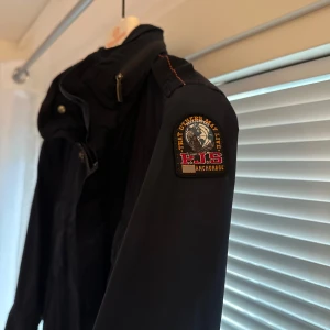 Svart vindjacka från Parajumpers - Säljer en svart vindjacka från Parajumpers med flera praktiska fickor framtill, dragkedja och knappar. Jackan har klassisk patch på ärmen och hög krage. Perfekt för dig som gillar stilren och funktionell design jättefin jacka men 2 små hål på baksidan syns knappt när man har den på sig mörkblå färg nypris 5000 kostar 500kr att laga hålen