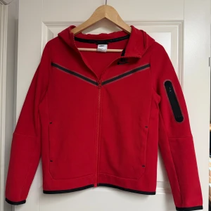 Röd Nike Tech hoodiejacka - Nike Tech hoodiejacka i rött med svarta detaljer och hel dragkedja framtill. Jackan har huva, lång ärm och en praktisk ficka med dragkedja på ena ärmen. Snygg V-formad detalj över bröstet och svarta kanter vid ärmslut och nederkant. Perfekt för en sportig look.