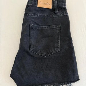 Svarta jeansshorts från Denim Day - Svarta jeansshorts från Denim Day med råa, fransiga benslut och klassisk femficksdesign. Shortsen har normal passform och knappgylf. Perfekta för en avslappnad stil.