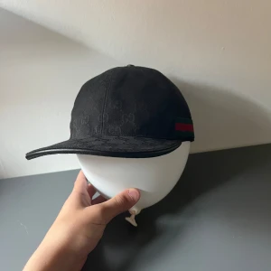 Svart keps från Gucci  - Snygg svart keps från Gucci med klassisk röd och grön rand på sidan. Kepsen har böjd skärm och justerbart spänne baktill. Perfekt accessoar för en trendig look.