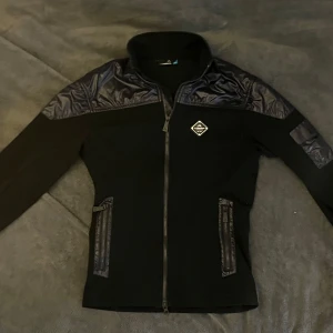 J.Lindeberg Cardigan - Svart jacka från J.Lindeberg med glansiga syntetdetaljer på axlar, ärmar och fickor. Dragkedja framtill och ståkrage. Två fickor med dragkedja och en liten patch på bröstet. Perfekt för en stilren look.