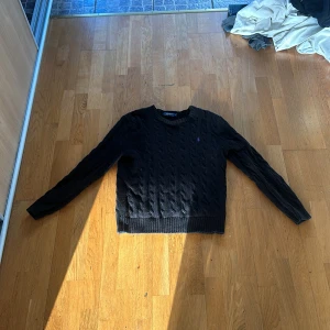 Svart stickad tröja från Polo Ralph Lauren - Kabelstickad svart tröja från Polo Ralph Lauren med rund halsringning och diskret broderad logga på bröstet. Klassisk modell med ribbade muddar vid ärmslut och nederkant. Perfekt för en stilren look.
