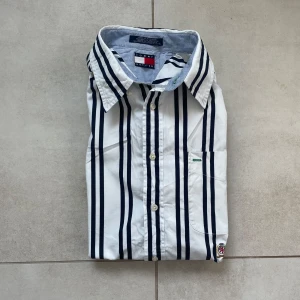 Vit och marinblå randig skjorta från Tommy Hilfiger - Vit skjorta med marinblå vertikala ränder från Tommy Hilfiger. Klassisk krage, knappstängning och lång ärm. Bröstficka med broderad detalj och logotyp. TommyHilfiger #VintageTommy #StripedShirt #MensFashion #PreppyStyle #TommyHilfigerShirt #VintageStyle #90sFashion #CasualWear #DesignerShirt #SmartCasual #SecondHandStyle #SustainableFashion #Menswear #ClassicLook