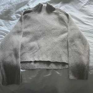 Beige ribbad stickad tröja från H&M - Säljer en beige stickad tröja från H&M med ribbad struktur och hög krage. Tröjan har långa ärmar och en något croppad passform. Perfekt för lager-på-lager och enkel att matcha med olika outfits.