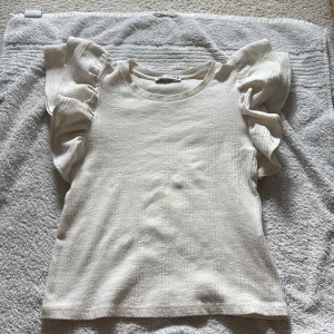 Vit ribbad topp med volangärmar från Zara - Säljer en vit ribbad topp från Zara med korta volangärmar. Toppen har rund halsringning och är tillverkad i ett mjukt material som ger en bekväm känsla. Perfekt för dig som vill ha en enkel men ändå trendig look.