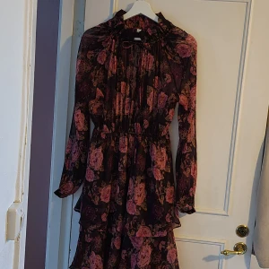 Blommig långklänning med volang från NA-KD - Romantisk långklänning med rosa och vinröda blommor på mörk botten. Klänningen har långa ärmar, volangdetaljer och knytning i halsen. Tillverkad i ett tunt, transparent polyestertyg som är just relativt transparent upptill. Nedtill är det dubbel med tyg. Aldrig använd.