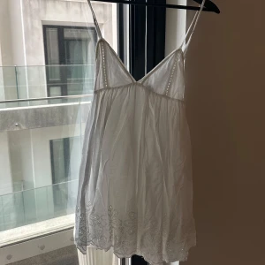 Vit somrig klänning med broderade detaljer - Söt vit klänning med smala axelband och v-ringning. Lätt och luftig modell med broderade blommor nertill och spetsdetaljer upptill. Perfekt för varma dagar eller sommarens alla tillfällen.