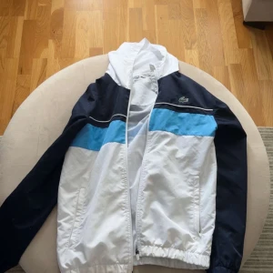 Vit och marinblå vindjacka från Lacoste - Snygg vindjacka från Lacoste i vitt och marinblått med ljusblå detalj över bröstet. Jackan har dragkedja framtill, ståkrage och två fickor. Perfekt för en sportig stil.