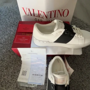 Valentino Garavani Open sneakers i vitt och svart - Säljer ett par snygga Valentino Garavani Open sneakers i vitt skinn med bred svart rem över mitten. Klassisk rund tå, platt sula och diskreta nitar baktill. Märkeslogga på plösen och häl. Perfekta för dig som gillar stilrena och exklusiva sneakers.