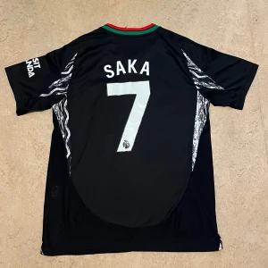 Arsenal svart bortatröja Saka 7 - Svart Arsenal bortatröja från Adidas med gröna och röda detaljer på axlarna. Tryck med Saka och nummer 7 på ryggen. Emirates Fly Better-logga på bröstet och klubbmärke i rött. Kortärmad modell med grafiskt mönster längs sidorna.