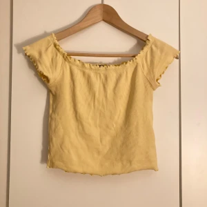 Gul ribbad offshoulder topp från Gina Tricot - Säljer en söt gul ribbad topp från Gina Tricot med offshoulder-modell och volangkant vid halsringningen och ärmslut. Toppen är croppad och har en figurnära passform, perfekt för vår och sommar🍋💞