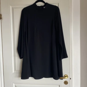 Svart långärmad klänning från Gina Tricot - En svart, enkel och stilren klänning från Gina Tricot med lång ärm och rak passform. Klänningen har hög hals och är tillverkad i ett mjukt, slätt material. Perfekt för dig som gillar minimalistisk stil.