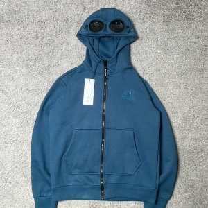 Blå hoodie från C.P Company - Säljer en helt ny blå hoodie från C.P. Company med dragkedja och de ikoniska glasögonen i huvan. Tröjan har broderad logga på bröstet och en stor magficka framtill. Perfekt för dig som gillar streetwear och unika detaljer. Hör av er vid frågor!