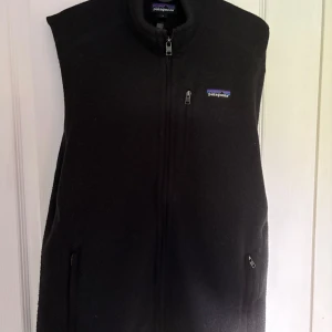 Patagonia väst - Säljer en svart fleeceväst från Patagonia i storlek M. Västen har dragkedja framtill och två fickor med dragkedja. Tillverkad av 100% återvunnen polyester och har en klassisk, enkel design med liten logotyp på bröstet.