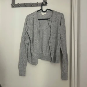 Ljusgrå kofta från H&M - En enkel och stilren ljusgrå kofta från H&M. Koftan har långa ärmar, rund halsringning och knäppning framtill. Perfekt att slänga över en topp eller klänning för en avslappnad look.