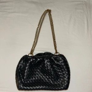 Svart flätad axelväska med guldkedja - Säljer en svart axelväska med snyggt flätat mönster och en rejäl guldfärgad kedja som axelrem. .