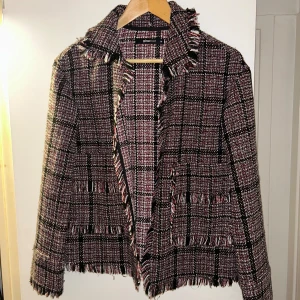 Rutig kavajjacka med fransar från Gina Tricot - Snygg rutig kavajjacka i svart, vit och röd med fransade kanter. Jackan har två fickor framtill och är öppen utan knäppning. Perfekt för lager på lager under höst och vår.