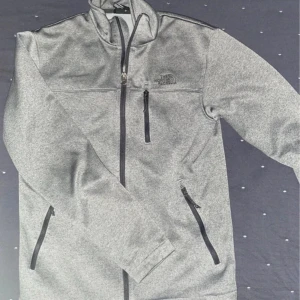 Grå zip-kofta från The North Face - Säljer en grå zip-kofta från The North Face med tre fickor med dragkedja och hög krage. Perfekt för lager-på-lager och enkel att matcha med det mesta. Diskret logga på bröstet och mjukt material.