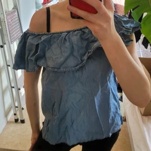 Blå jeans offshoulder volangtopp - Säljer en ljusblå jeans offshoulder topp med volangdetalj längs överdelen. Toppen är ärmlös och har en luftig passform, perfekt för varma dagar. Snygg att matcha med både jeans och kjol! Så himla mycket och trendig nu i sommar!