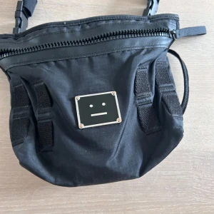 Svart axelväska från Acne Studios - Säljer en svart axelväska från Acne Studios med justerbar rem och dragkedja upptill. Väskan har det klassiska Acne Studios-face-loggan framtill och meshficka på baksidan. Perfekt för att bära det viktigaste med stil.