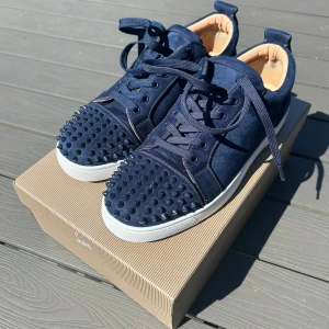 Mörkblå sneakers med nitar från Christian Louboutin - Säljer ett par mörkblå sneakers från Christian Louboutin i mocka med rund tå och snörning. Skorna har svarta nitar på tån och sidorna samt den klassiska röda sulan. Perfekta för dig som vill sticka ut med en lyxig och edgy stil. Pris kan diskuteras 