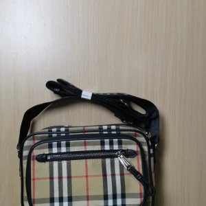 Rutig axelväska från Burberry - Säljer en klassisk axelväska Burberry med det ikoniska rutiga mönstret i beige, svart, rött och vitt. Väskan har en justerbar axelrem och en praktisk dragkedja framtill. Perfekt inför sommar. Insta👉 märke _kläder 