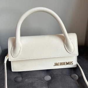 Vit handväska - Säljer en  vit handväska med guldfärgad logga och detaljer. Väskan har ett avtagbart axelband och ett stadigt handtag. Axelbandet har gått sönder men går att laga. Köpt i Spanien