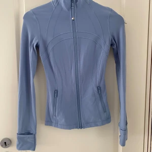 Ljusblå träningsjacka från Lululemon - Säljer en ljusblå träningsjacka från Lululemon med figurnära passform. Det är Nulu lång i storlek 0! 