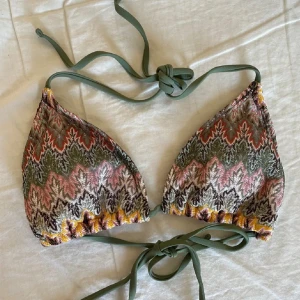 Mönstrad trekantsbikini med snörning - Säljer en snygg trekantsbikini med knytband i grönt och ett färgglatt mönster i grönt, orange, rosa och gult. Bikinin har snäckformad detalj i mitten och är perfekt för sommaren. Materialet känns syntetiskt och toppen har normal passform. Väldigt lik den på första bilden men inte precis samma! 