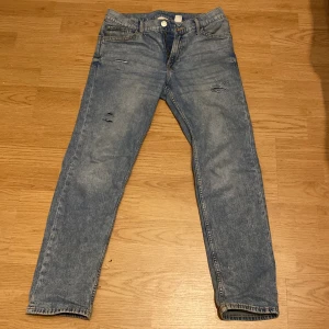 Blå slim fit jeans från H&M - Säljer ett par blå slim fit jeans från H&M med justerbar midja. Jeansen har slitna detaljer framtill och klassisk femficksdesign. Perfekta för dig som gillar en smalare passform och enkel stil.