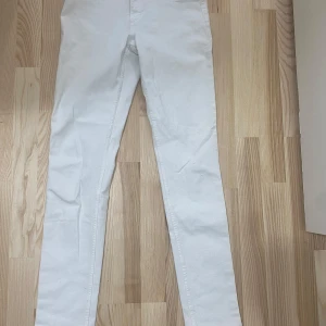 Vita skinny jeans - Säljer ett par vita skinny fit jeans i denim med klassisk femficksdesign och normalhög midja. Byxorna har dragkedja och knapp framtill och är perfekta för en stilren look.