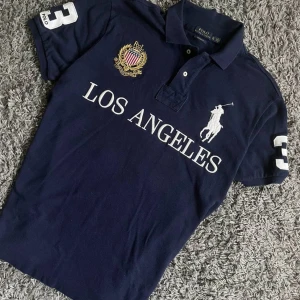 Mörkblå pikétröja från Polo Ralph Lauren - Säljer en mörkblå pikétröja från Polo Ralph Lauren med broderad logga, stort tryck 'LOS ANGELES' och siffran 3 på ärmarna. Klassisk krage och knappar framtill. Perfekt för en sportig och stilren look.