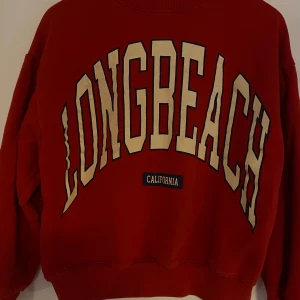 Röd sweatshirt med tryck från Pull&Bear - Röd sweatshirt från Pull&Bear med stort vitt och svart 'LONG BEACH CALIFORNIA'-tryck på framsidan. Klassisk rund halsringning och ribbade muddar vid ärmslut och nederkant. Perfekt för en avslappnad och sportig stil.
