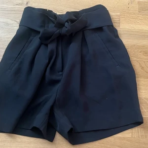 Svarta shorts med knytband från H&M - Stilrena svarta shorts från H&M med hög midja och knytband i midjan. Shortsen har två fickor fram och dekorativa fickor bak. Perfekta för en trendig och bekväm look.