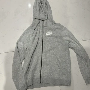 Grå hoodie från Nike - Säljer en grå hoodie från Nike med dragkedja och huva. Tröjan har långa ärmar, två fickor framtill och Nike-logga i vitt på bröstet. Perfekt för en avslappnad stil.
