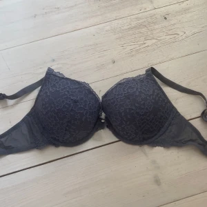 Mörkgrå spets-bh - Säljer en mörkgrå bh med vacker spetsdetalj över hela kuporna. Bh:n har vadderade kupor och justerbara axelband för bästa passform. Perfekt för dig som vill ha något både snyggt och bekvämt.