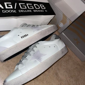 Vita sneakers från Golden Goose Superstar - Säljer ett par vita Golden Goose Superstar sneakers med grå snörning och silverdetaljer. Skorna har den klassiska stjärnan på sidan och GGDB-logga. Perfekta för dig som gillar stilrena och trendiga sneakers.