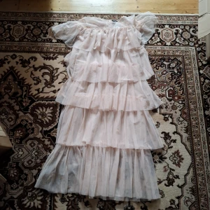 Ljus volangklänning med offshoulder - Romantisk ljus långklänning med flera lager volanger och offshoulder-design. Klänningen har en luftig och drömmig känsla, perfekt för sommarens speciella tillfällen. Tillverkad i ett tunt, transparent material som ger ett mjukt fall.