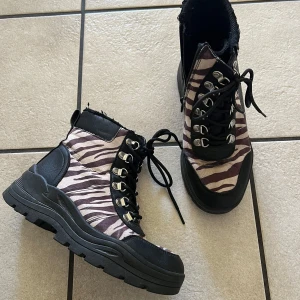 Zebramönstrade boots med grov sula - Säljer ett par boots med zebramönster i svart och beige. Skorna har grov svart sula, snörning med metallhål och svarta detaljer vid häl och tå. Ovandelen är i syntetmaterial och de har en cool, edgy look.