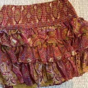 Mönstrad volangkjol från Zara - Färgglad kortkjol från Zara med paisleymönster i rött, gult och rosa. Kjolen har flera volanger och smock i midjan för en bekväm passform. Perfekt för dig som gillar boho-stil och vill ha något unikt i garderoben.