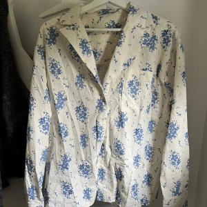 Vit blommig skjorta från NA-KD - Säljer en vit skjorta från NA-KD med blått blommigt mönster. Skjortan har lång ärm, klassisk krage och en bröstficka. Tillverkad i ett lätt och luftigt material, perfekt för vår och sommar.