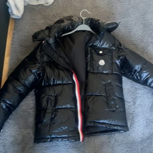 Svart pufferjacka från Moncler - Säljer en svart pufferjacka från Moncler med huva och dragkedja framtill. Jackan har en ficka med knapp på bröstet och Moncler-logga. Klassisk glansig finish och quiltad design.