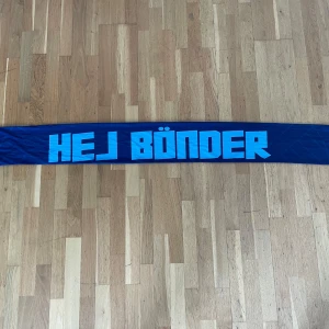 ”Hej bönder vi e DIF” -halsduk från Järnkaminerna - Säljer en limiterad blå halsduk från Järnkaminerna med texten 'HEJ BÖNDER' på ena sidan och 'VI É DIF!' på andra. Halsduken har JK-sköldar ton-i-ton i bakgrunden och blå fransar i ändarna. 