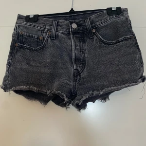 Svarta Levi's jeansshorts - Snygga svarta jeansshorts från Levi's med råa fransade kanter och klassisk femficksdesign. Shortsen är midwaist och har slitna detaljer för en cool vintagekänsla. Perfekta för sommardagar och festivaler.