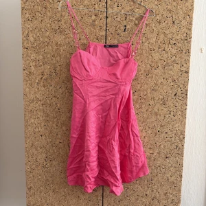 Rosa kort klänning från Zara - Supersöt rosa kort klänning från Zara med tunna axelband och coola plastdetaljer i axelbanden. Klänningen har bystformade kupor och lätt utsvängd kjol. Perfekt för dig som vill sticka ut med en färgglad och trendig look.