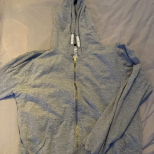 Grå hoodie med dragkedja från MARCY - Grå hoodie från MARCY med broderad logga på bröstet. Tröjan har huva med snörning, hel dragkedja framtill och två fickor. Tillverkad i mjuk bomullsmix, perfekt för chill dagar. Långärmad och klassisk passform.