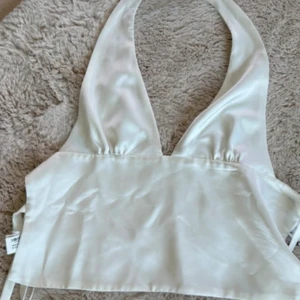 Vit halterneck topp från Zara - Snygg vit halterneck topp från Zara i glansigt satinliknande material. Toppen har djup v-ringning och knytning i ryggen för en justerbar passform. 
