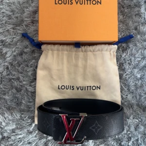 Louis Vuitton svart skärp med LV-spänne - Snyggt svart Louis Vuitton-bälte i skinn med klassiskt monogrammönster och ett stort LV-spänne i silver och rött. Bältet har en stilren look och levereras med originalpåse och box. Perfekt accessoar för att lyfta din outfit.