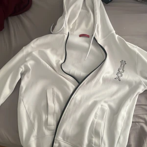 Vit hoodiejacka från Hugo Boss - Snygg vit hoodiejacka från Hugo Boss med svart logotyptryck både fram och bak. Jackan har dragkedja, huva med snörning och två fickor framtill. Materialet är mjukt och känns sportigt, perfekt för en chill och stilren look.