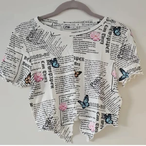 Vit croppad topp med tidningsmönster FB Sister - Supercool vit croppad topp från FB Sister med tidningsinspirerat print, fjärilar och rosa blommor. Toppen har korta ärmar, vågig nederkant och är tillverkad i mjuk viskosblandning med stretch. Perfekt för dig som vill sticka ut med en unik och trendig look.
